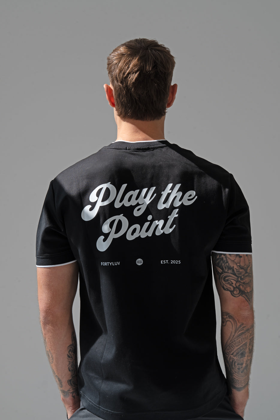 Person wearing a padel black t-shirt with 'Play the Point' text on the back against a gray background